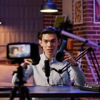 Asian influencer filming headphones review on podcast episode, recording recommendation video on camera. Live broadcasting talk show with wireless headset product on social media.